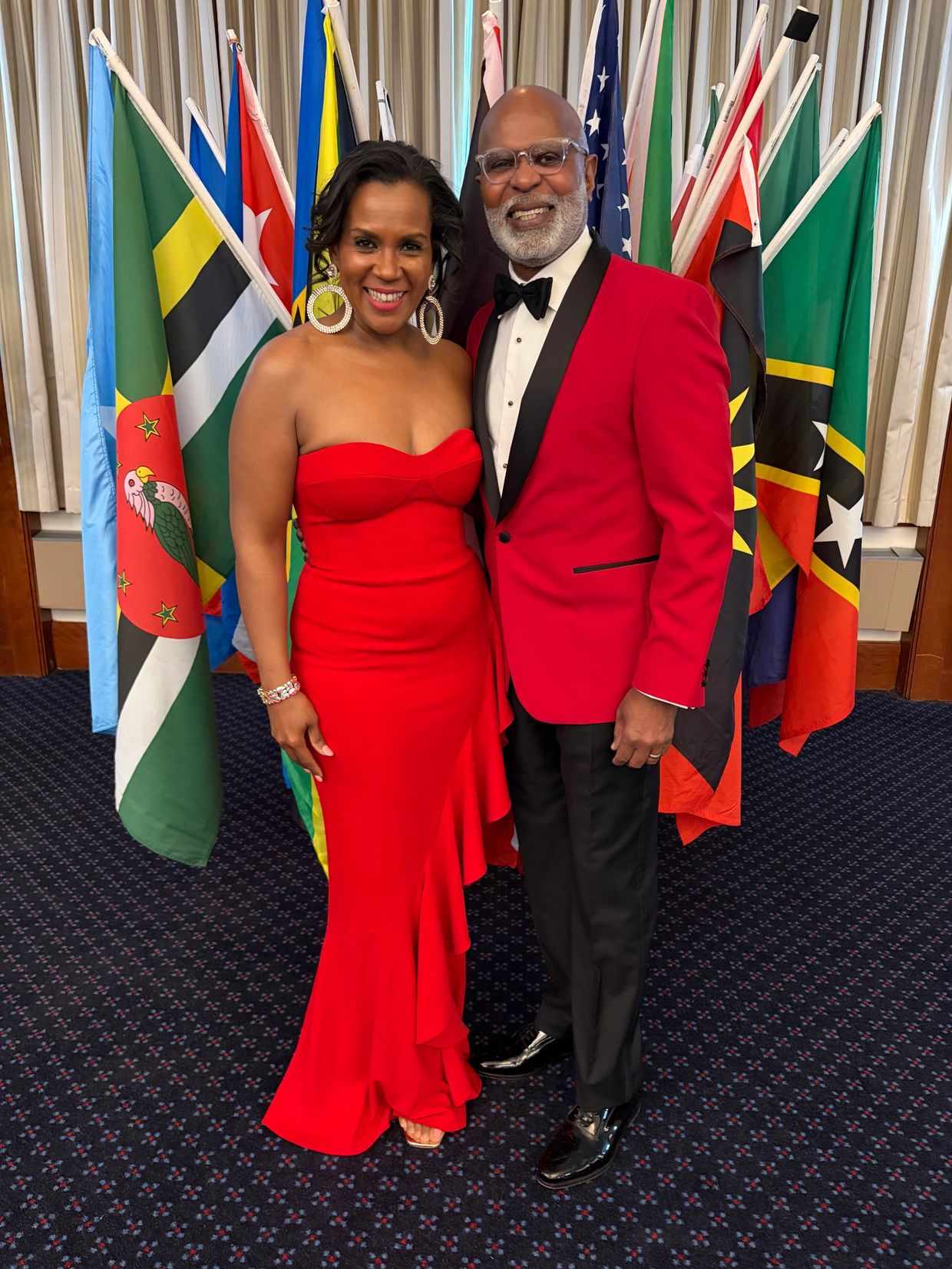A couple dressed elegantly in red stands smiling in front of international flags.
