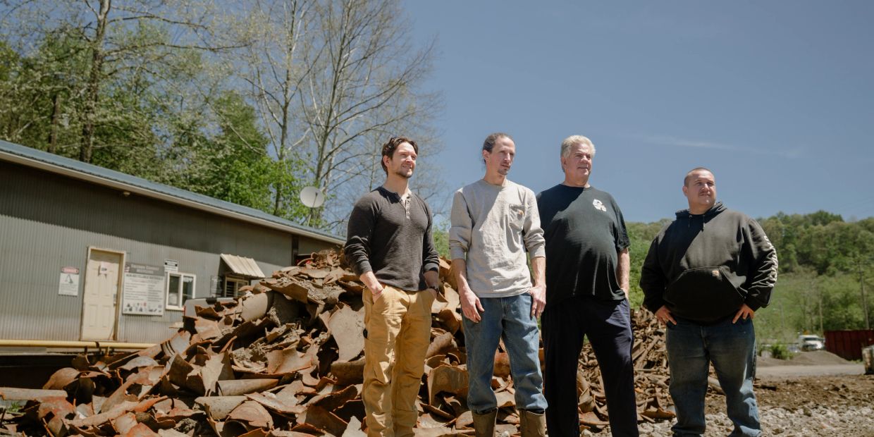Four men standing outdoors near a pile of rusted metal.