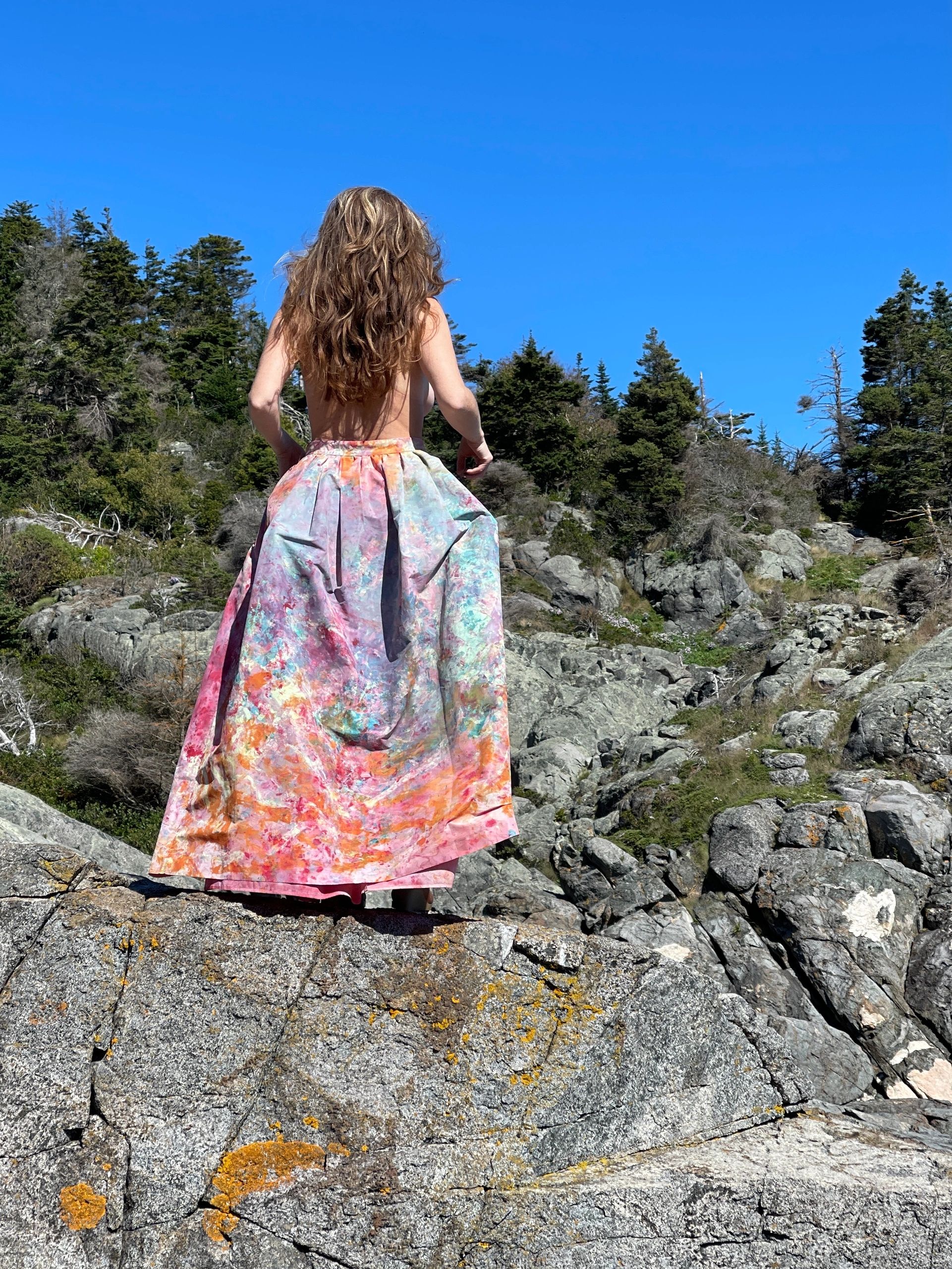 Woman with flowing hair in a colorful skirt stands on rocky terrain under clear blue sky.
