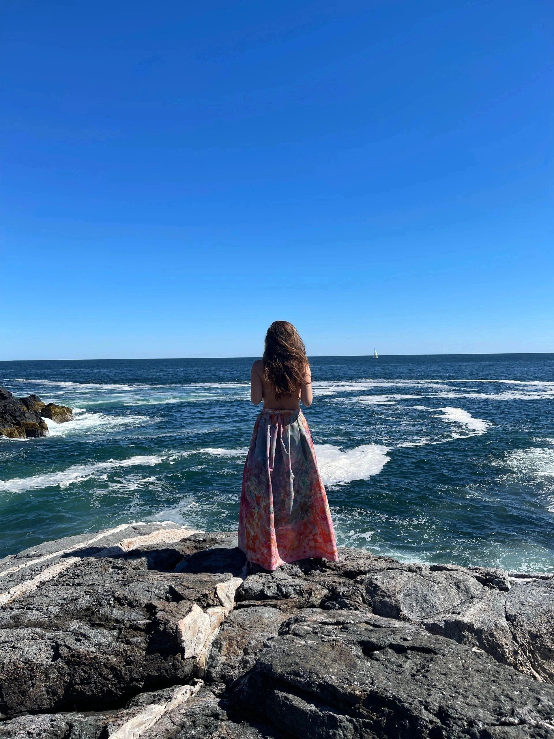 A woman in a colorful skirt stands on rocks, gazing at the vast ocean under a clear blue sky.