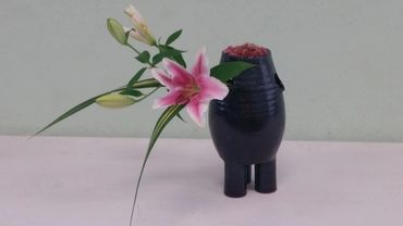 A pink lily and green buds arranged in a black vase on a white surface.
