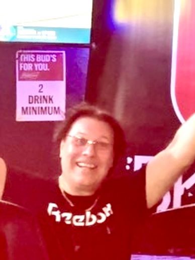 Smiling person raising arms in a bar with a '2 drink minimum' sign.