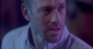 still image of man in purple-blue lighting, looking quizzically at something out of frame