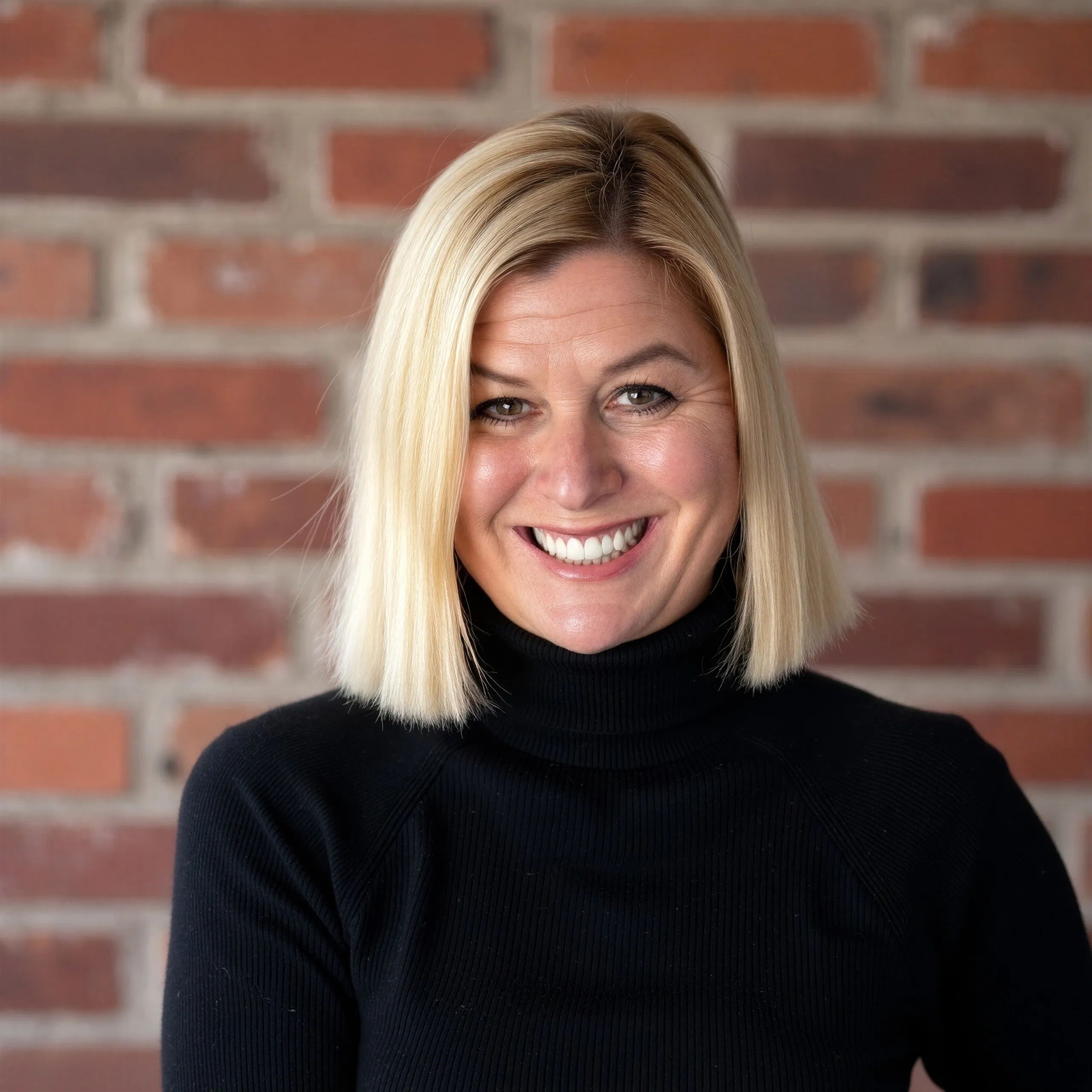 Smiling blonde woman with a black turtleneck against a brick wall.