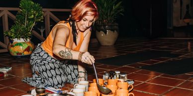 Red-haired woman in an orange top pouring cacao into orange coffee cups on a tiled outdoor deck