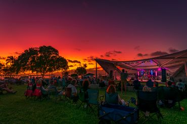 People relaxing on the grass at an outdoor concert during a vibrant sunset.