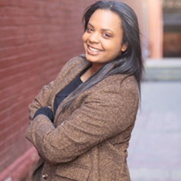 Smiling woman in a brown jacket standing with arms crossed outdoors.