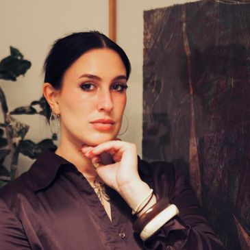 A woman with dark hair and bracelets poses thoughtfully indoors.