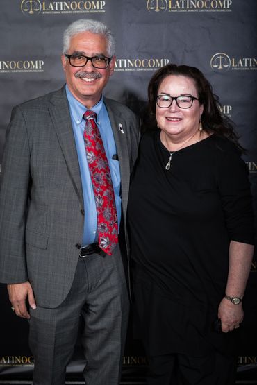Two people smiling at a LatinoComp event with a branded backdrop.