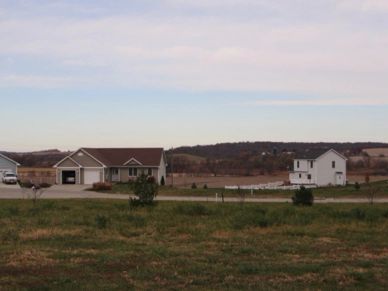 Suburban houses with open fields and a distant tree line under a cloudy sky.