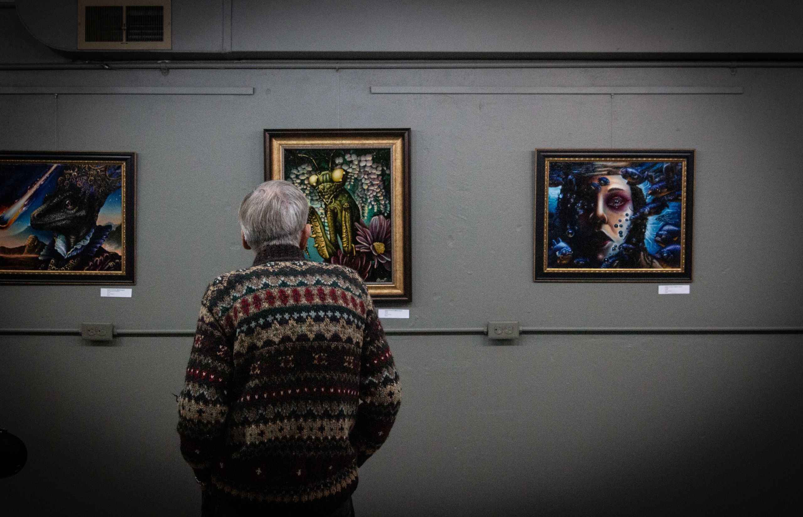 Man in patterned sweater observing surreal paintings in art gallery.