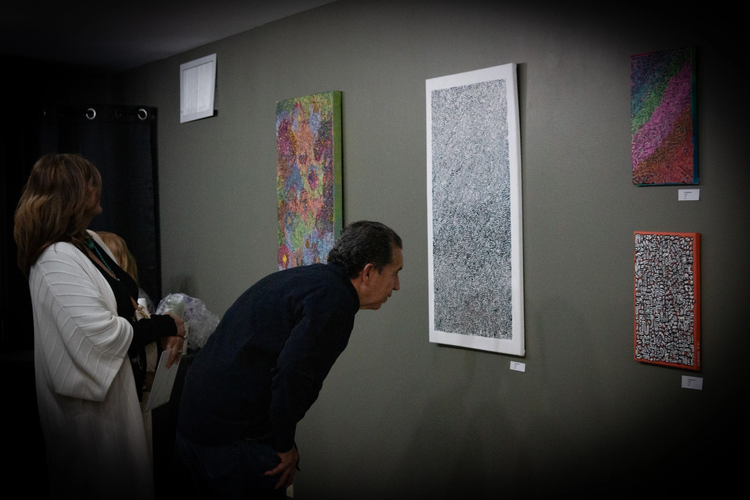 Two people closely examining abstract paintings on a gallery wall.
