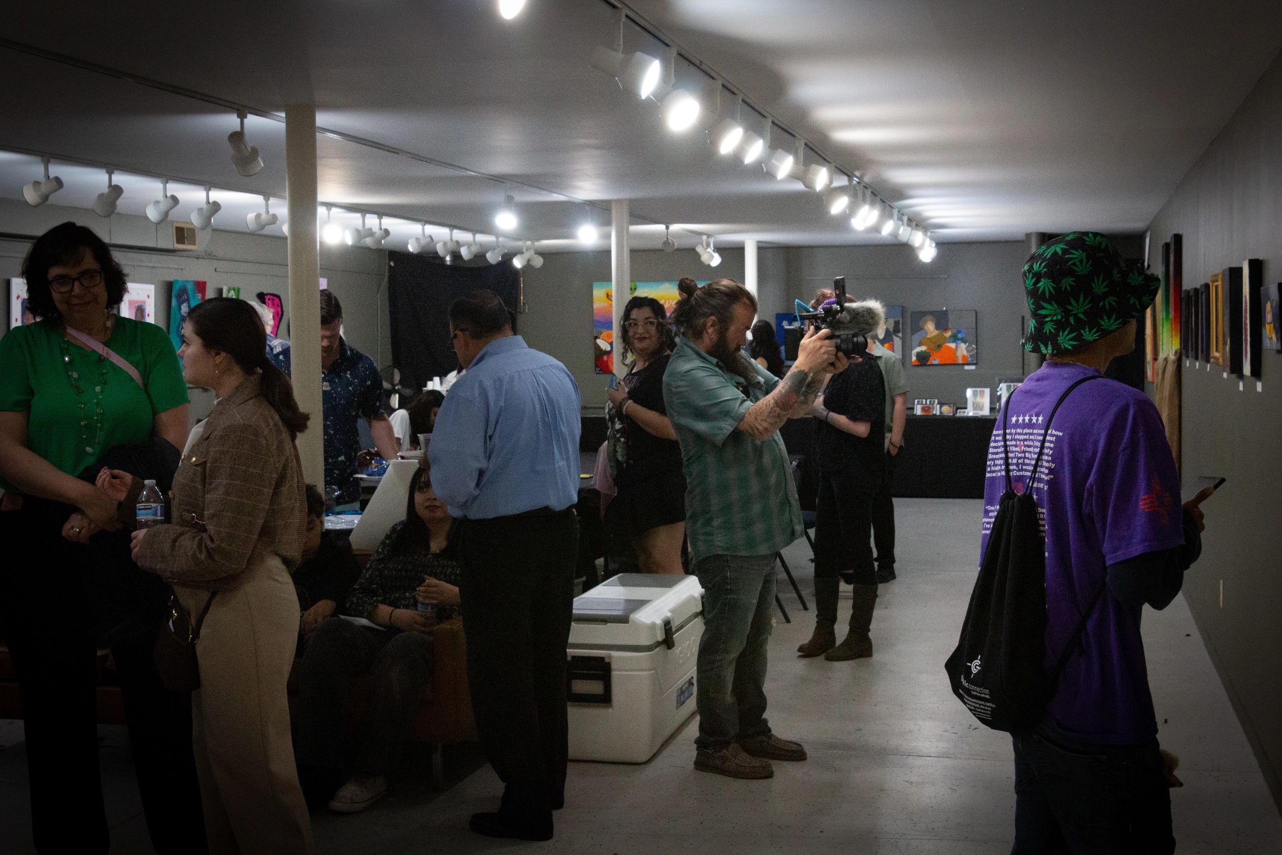 People mingling and viewing art in a gallery with a videographer capturing moments.