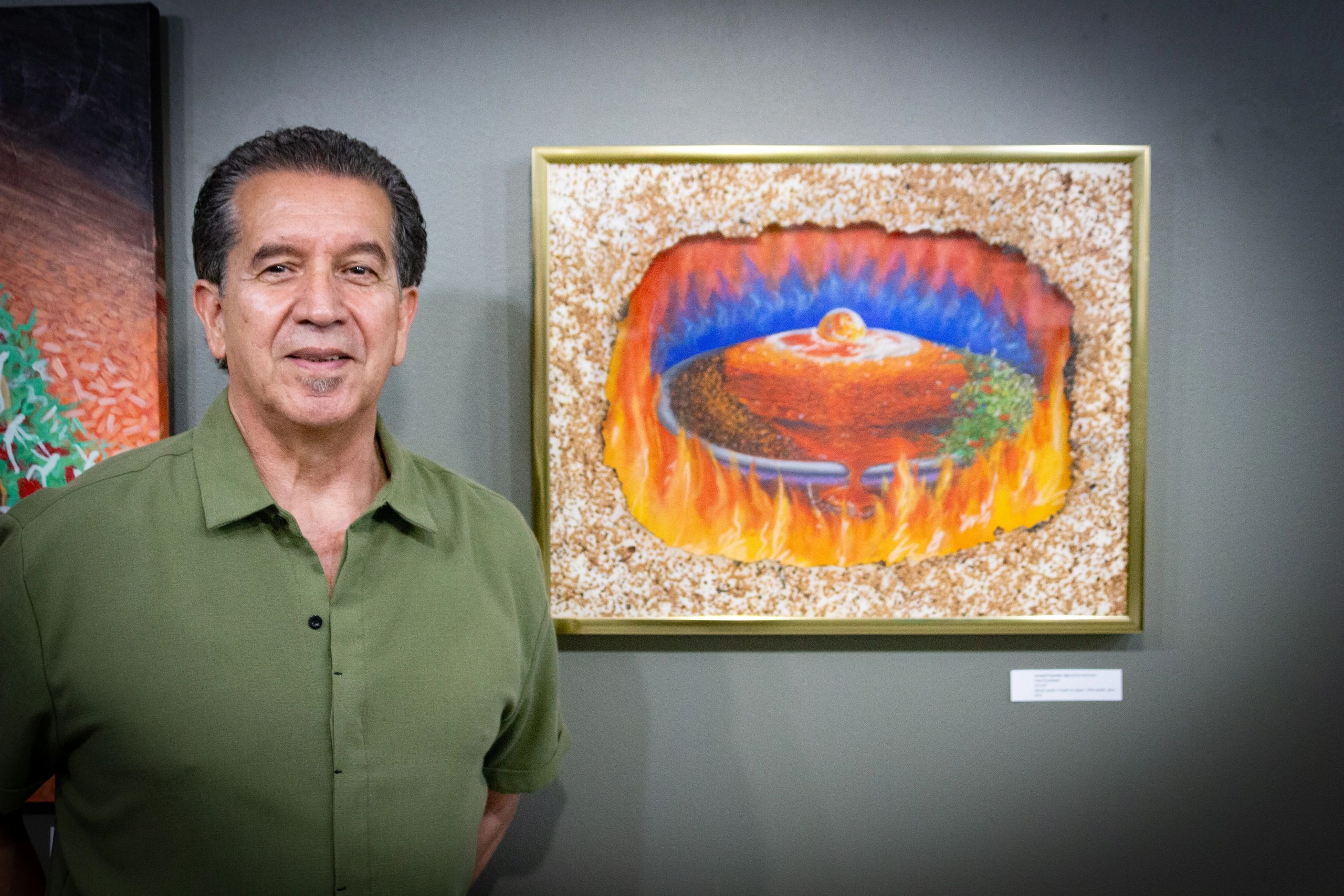 Man in green shirt standing next to a colorful framed painting with flames.