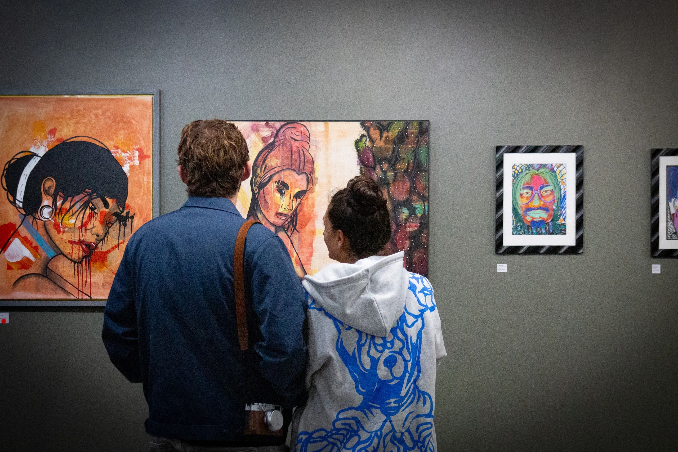 Two people observe colorful portrait paintings in an art gallery.