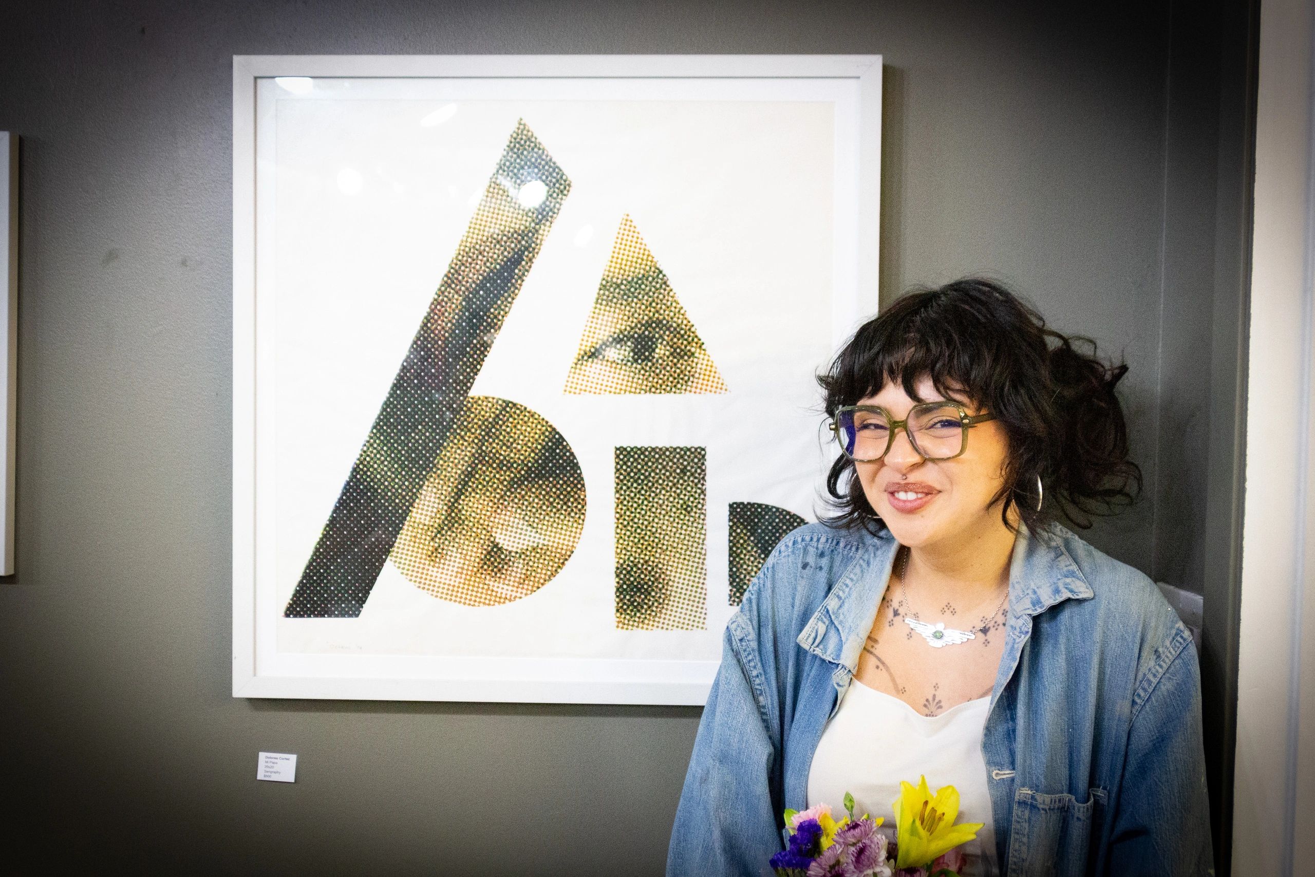 A smiling woman with glasses and flowers poses in front of framed art.