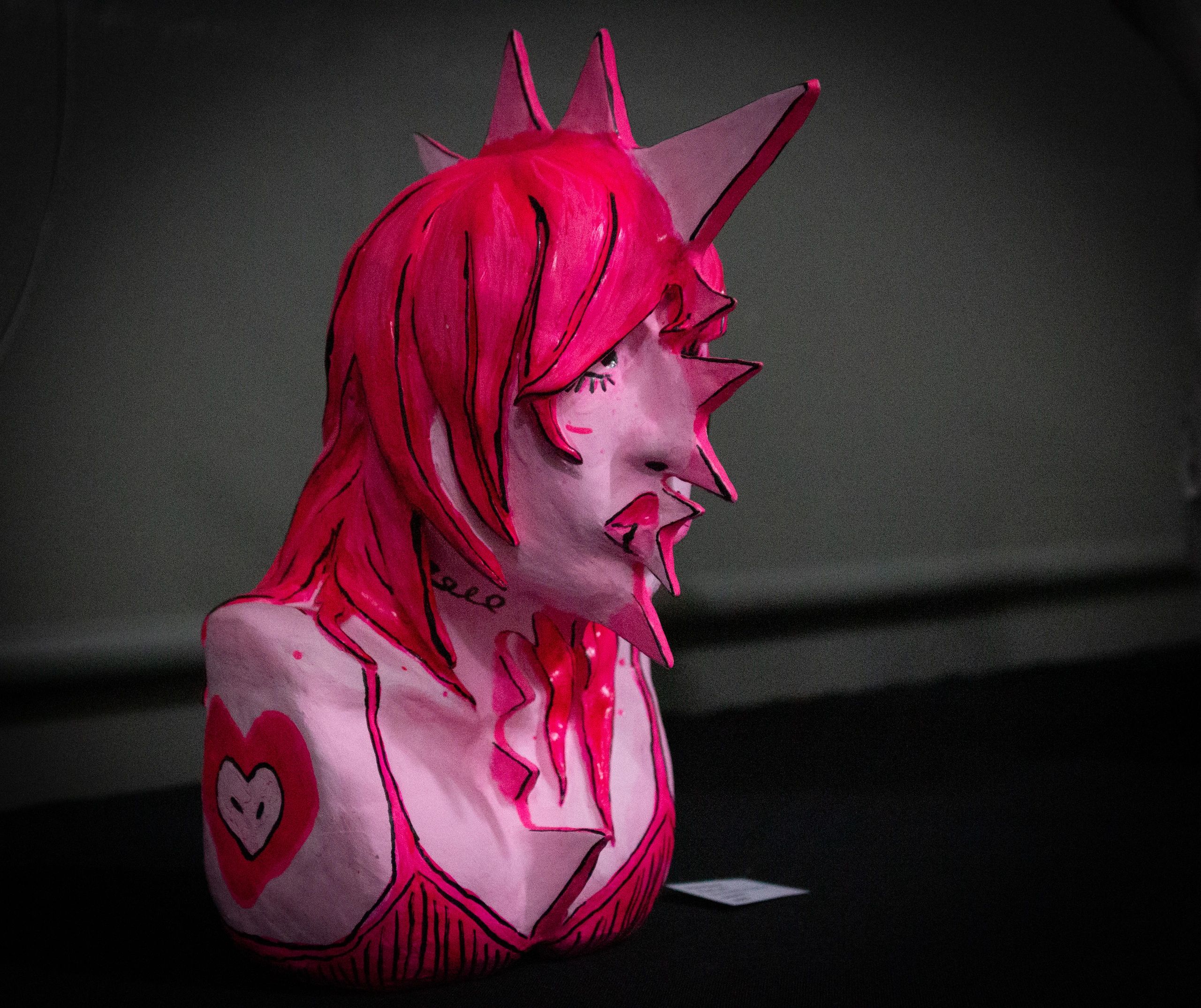 Pink sculptural figure with spikes and heart designs on a dark background.