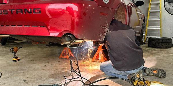 Person welding underneath a red Mustang car on jack stands in a garage.