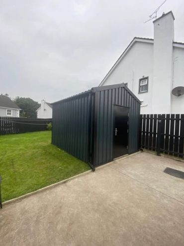A sleek black metal shed in a backyard with a concrete driveway and green lawn.