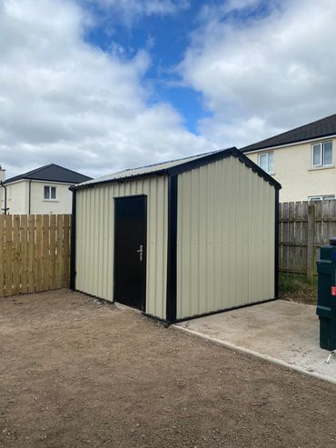 A beige metal garden shed with a black door in a fenced backyard.