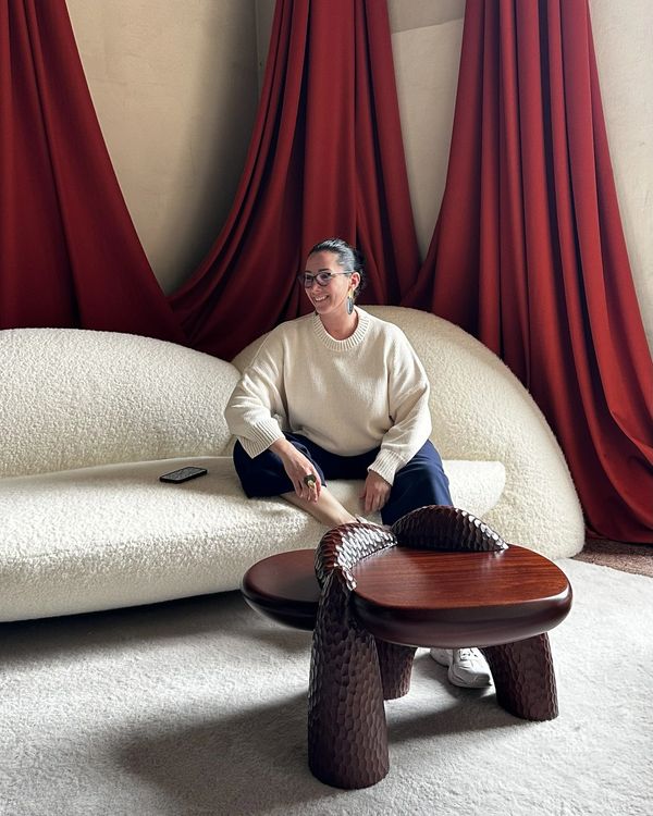 A woman sitting on a white textured couch with red curtains and a unique wooden table.