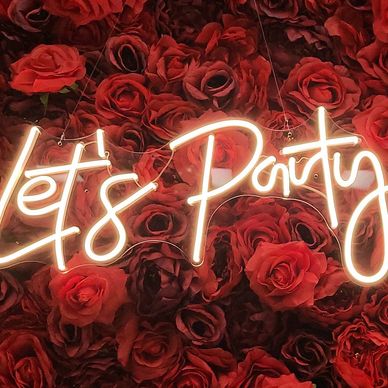 Neon sign reading "Let's Party" over a wall of red roses.