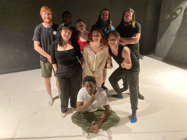 A diverse group of nine young adults posing together on a white floor with a dark background.