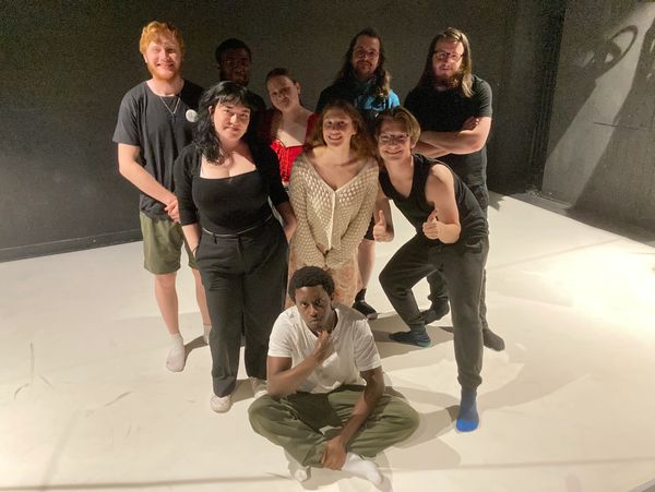 A diverse group of nine young adults posing together on a white floor with a dark background.