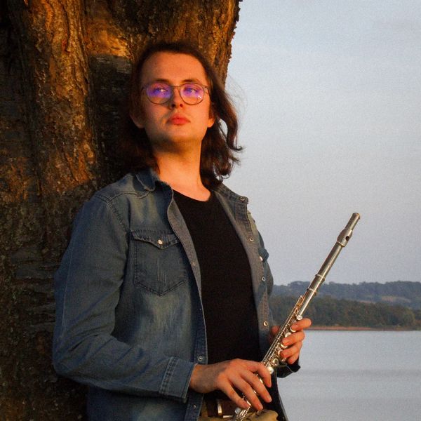 Person with glasses holding a flute, standing by a tree near water.