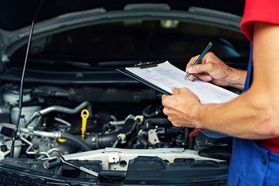 Mechanic inspecting car engine and taking notes on clipboard.