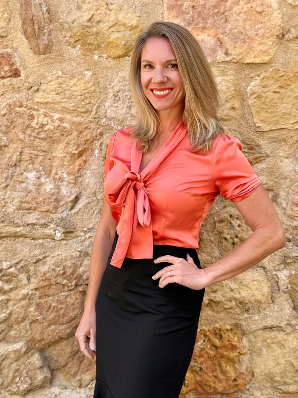 Mujer sonriente con blusa naranja y falda negra frente a pared de piedra.