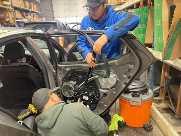 Two people are in the process of replacing a car door glass. One person is holding the glass up