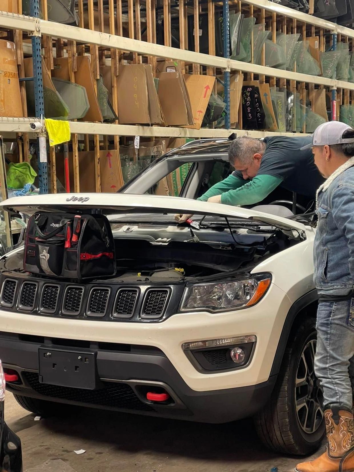 1 person replacing a car's front  windshield and second person with boots looking at how he works