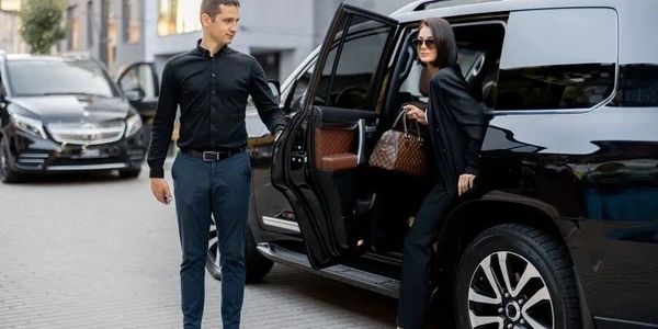 A man opens a car door for a woman dressed in black with sunglasses and a handbag.