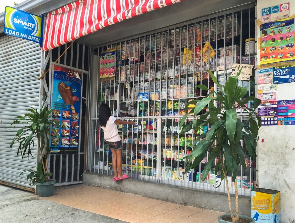 A girl stands on a ledge reaching through bars at a small neighborhood store.