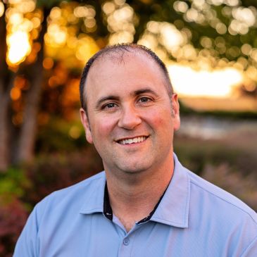 Man smiling outdoors with sunset background.