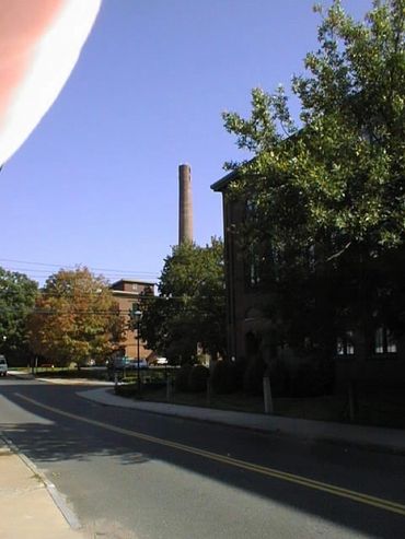 A quiet street with a tall chimney and trees on a sunny day.
