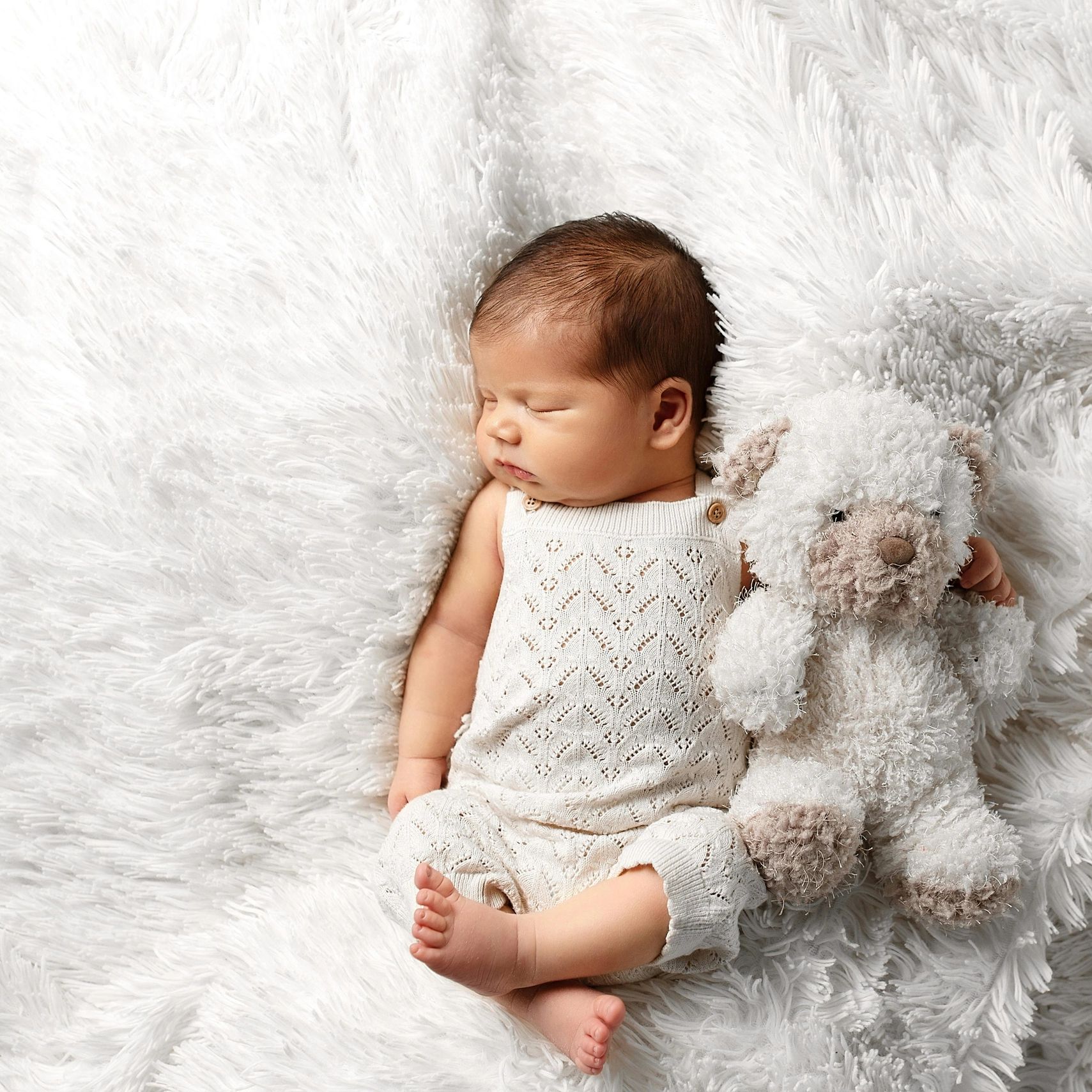 Sleeping newborn baby cuddling a soft teddy bear on a fluffy white blanket.