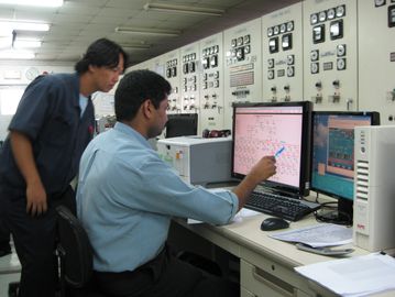 Two men working together in a control room with multiple monitors and control panels.