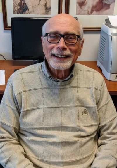 Elderly man smiling warmly, wearing glasses and a beige sweater, sitting indoors.