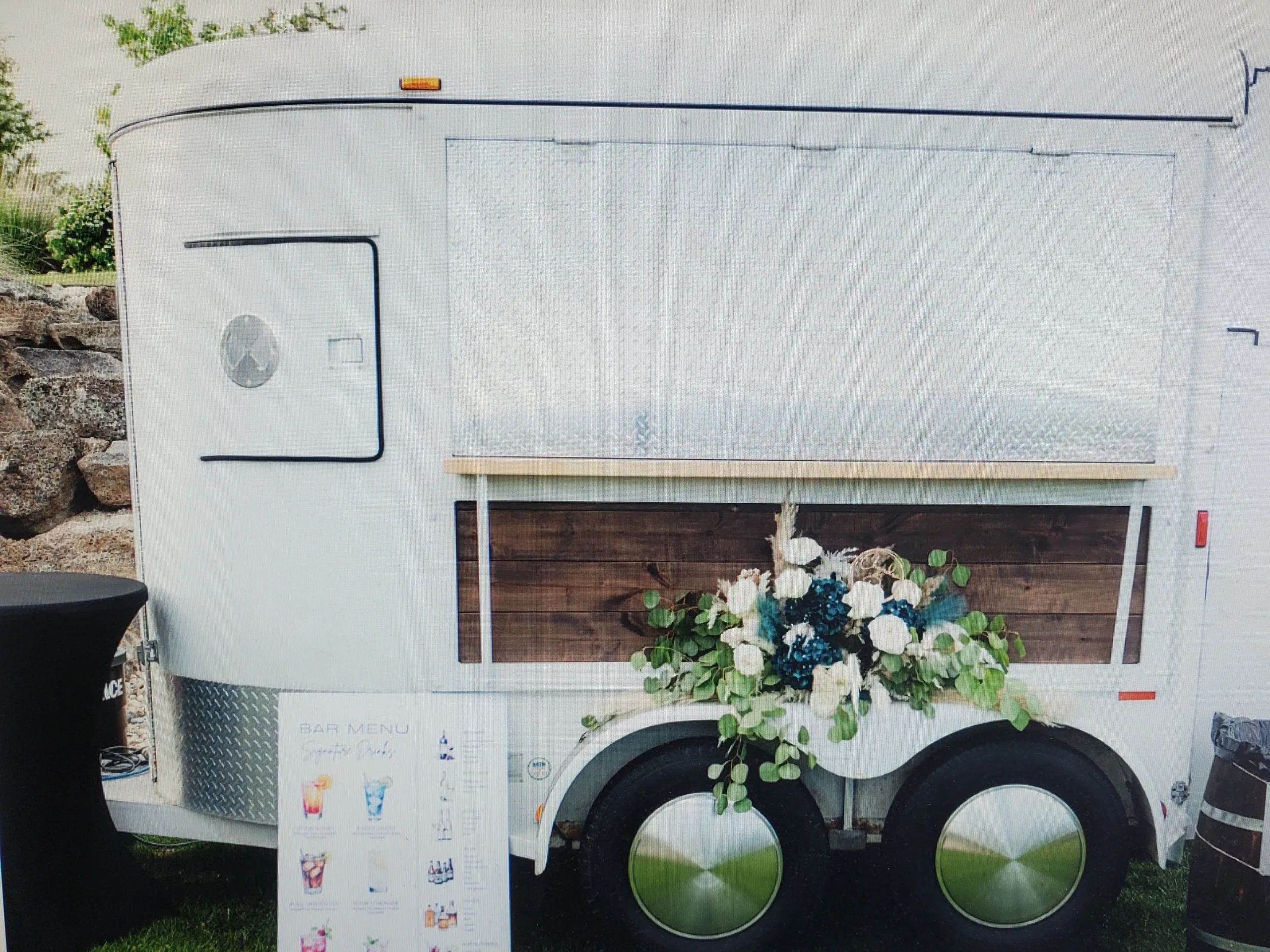 A white vintage trailer decorated with flowers and a bar menu sign.