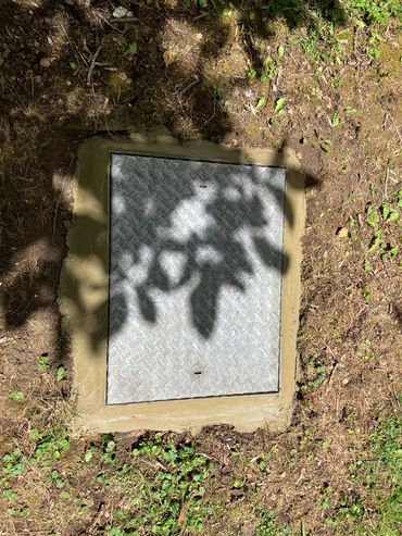 A metal hatch set in the ground surrounded by dirt and plants.