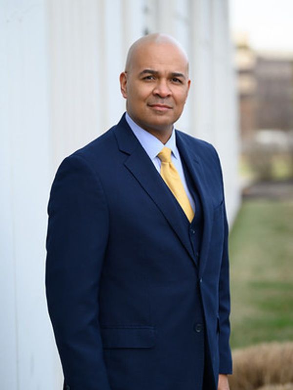 A man in a navy blue suit with a yellow tie stands outdoors.