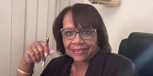 A woman with glasses holding a pen and smiling indoors.