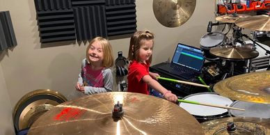 Two young girls in a music room with a drum set and cymbals.