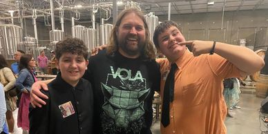 Three people smiling together inside an industrial brewery setting.