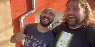 Two men smiling happily in front of a red brick wall.