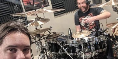 Two men posing with a drum set in a soundproof room, one giving a thumbs-up.