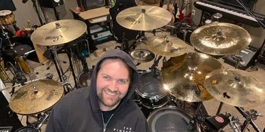 A smiling man in a hoodie surrounded by a drum set and musical equipment.