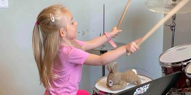 A young girl playing drums with a stuffed bunny on the drum.